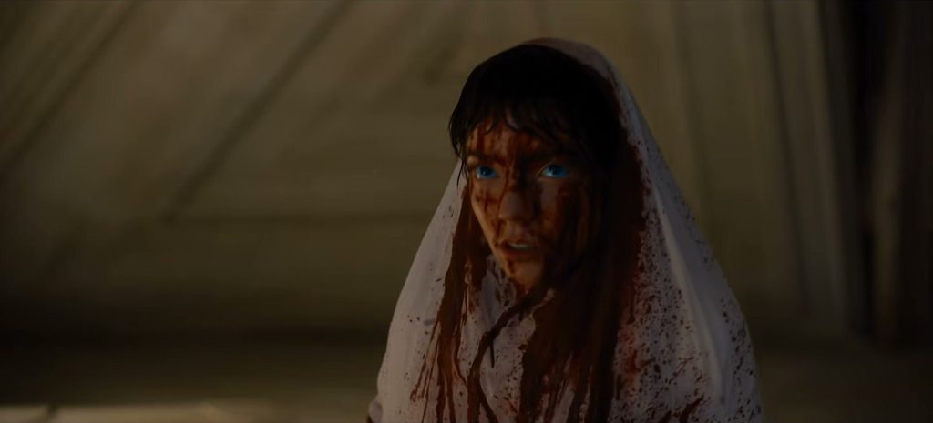 Une jeune femme aux yeux bleus, voile blanc sur la tête, le visage et les cheveux couverts de sang, dans une pièce sombre.