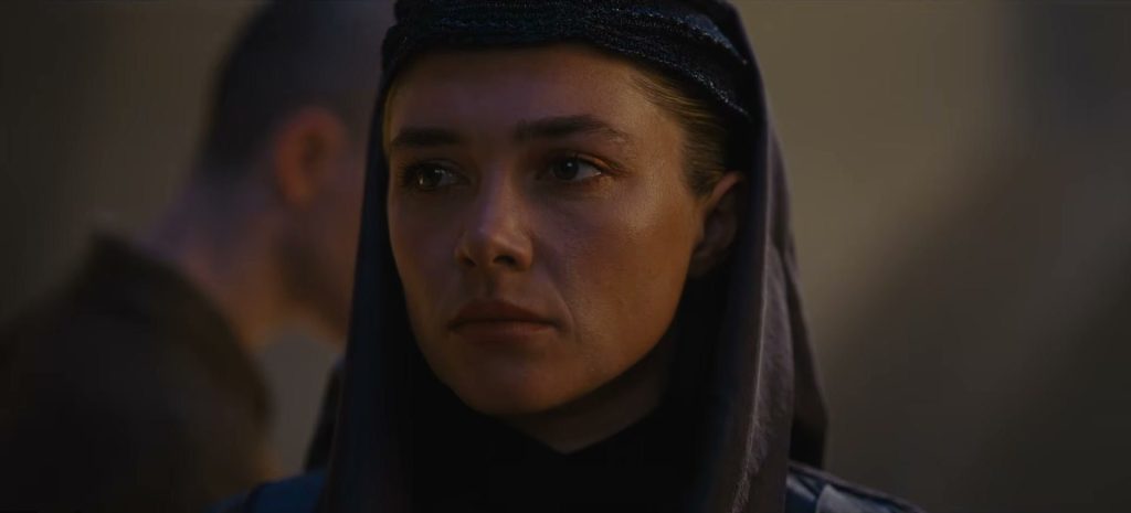 Florence Pugh en princesse Irulan, voile sombre sur la tête, le regard fixe dans une pièce sombre, une silhouette floue derrière elle.