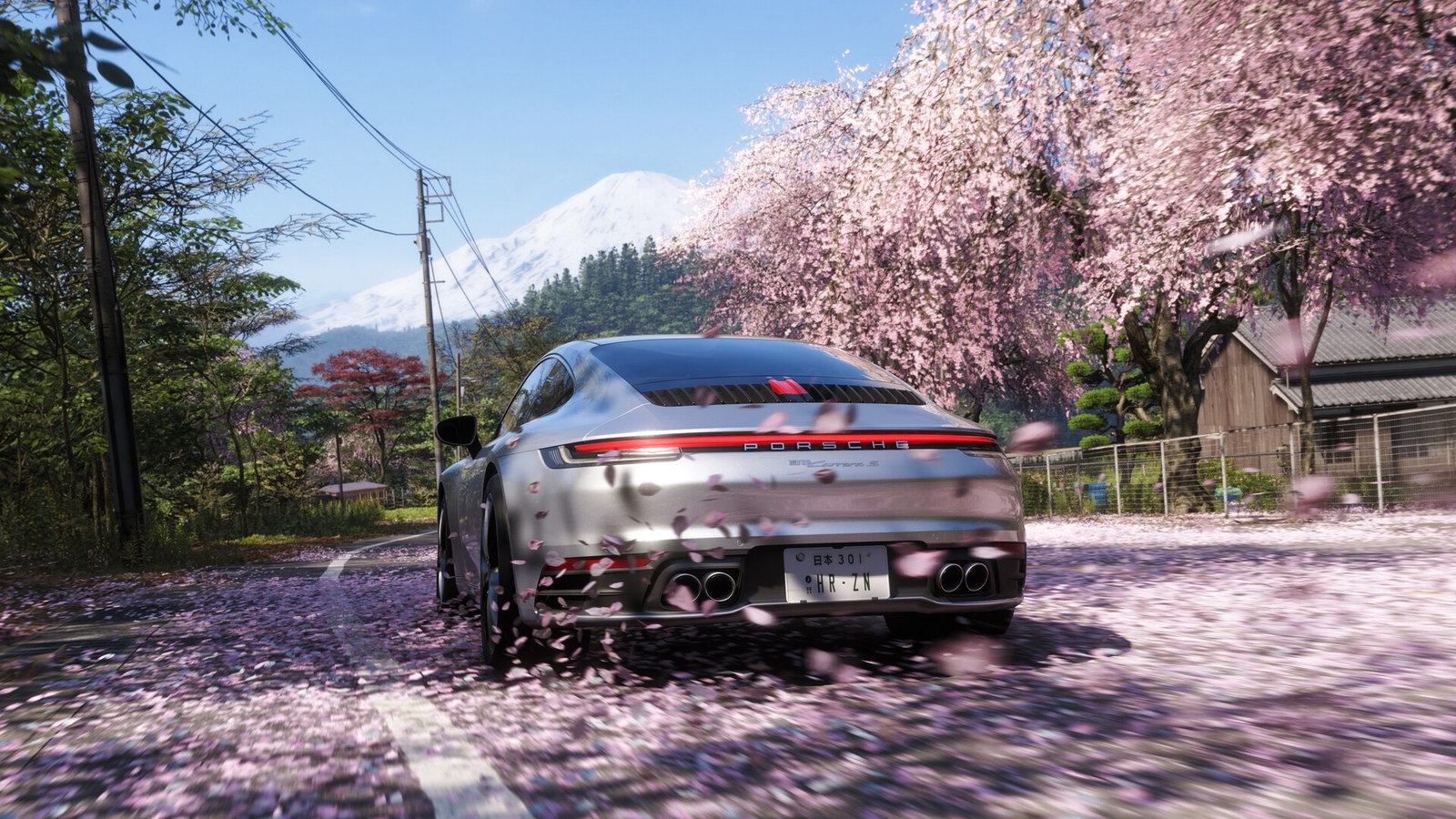 Une voiture de sport grise roule sur une route bordée de cerisiers en fleurs, une montagne enneigée au fond.