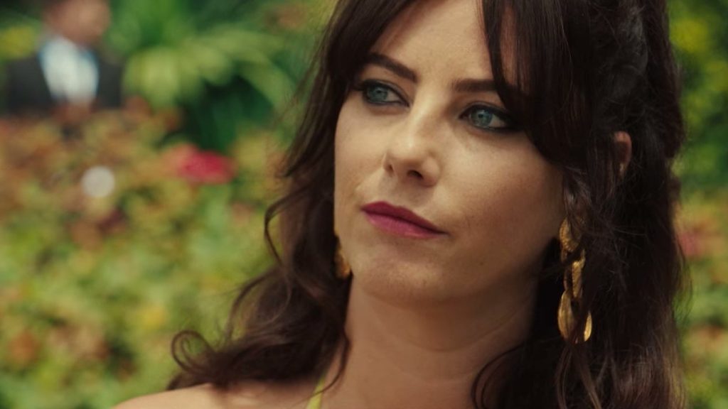 Kaya Scodelario en Susie Glass, cheveux bruns et boucles d’oreilles dorées, dans un jardin fleuri.