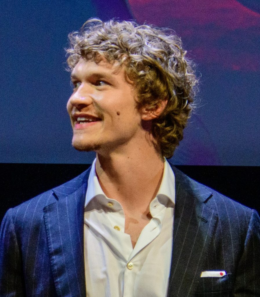 Connor Storrie, cheveux bouclés, veste bleu marine à fines rayures et chemise blanche ouverte, sourit en regardant sur sa droite, devant un fond bleu.