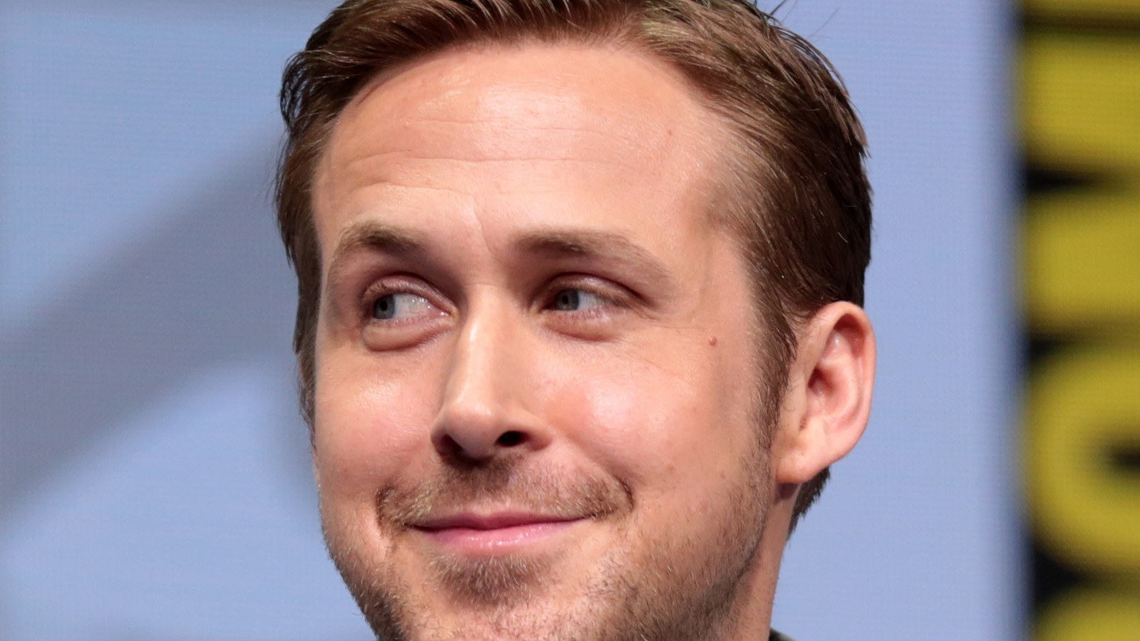 Ryan Gosling, cheveux courts, sourit en regardant sur le côté, devant un panneau jaune et noir du Comic-Con.