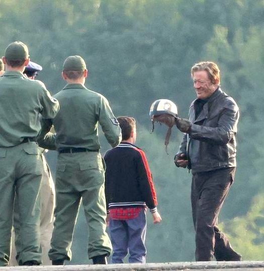 Un groupe d'hommes en uniformes verts et d'enfants en casquette, vus de dos, devant un homme en veste sombre qui tient un casque de course blanc.