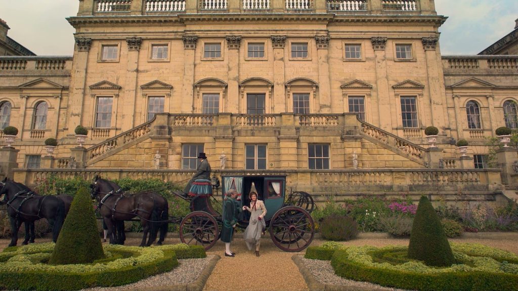 Une calèche attelée à quatre chevaux noirs s’arrête devant la façade de pierre blonde d’un grand château à balustrades, entre des buis taillés en cône.