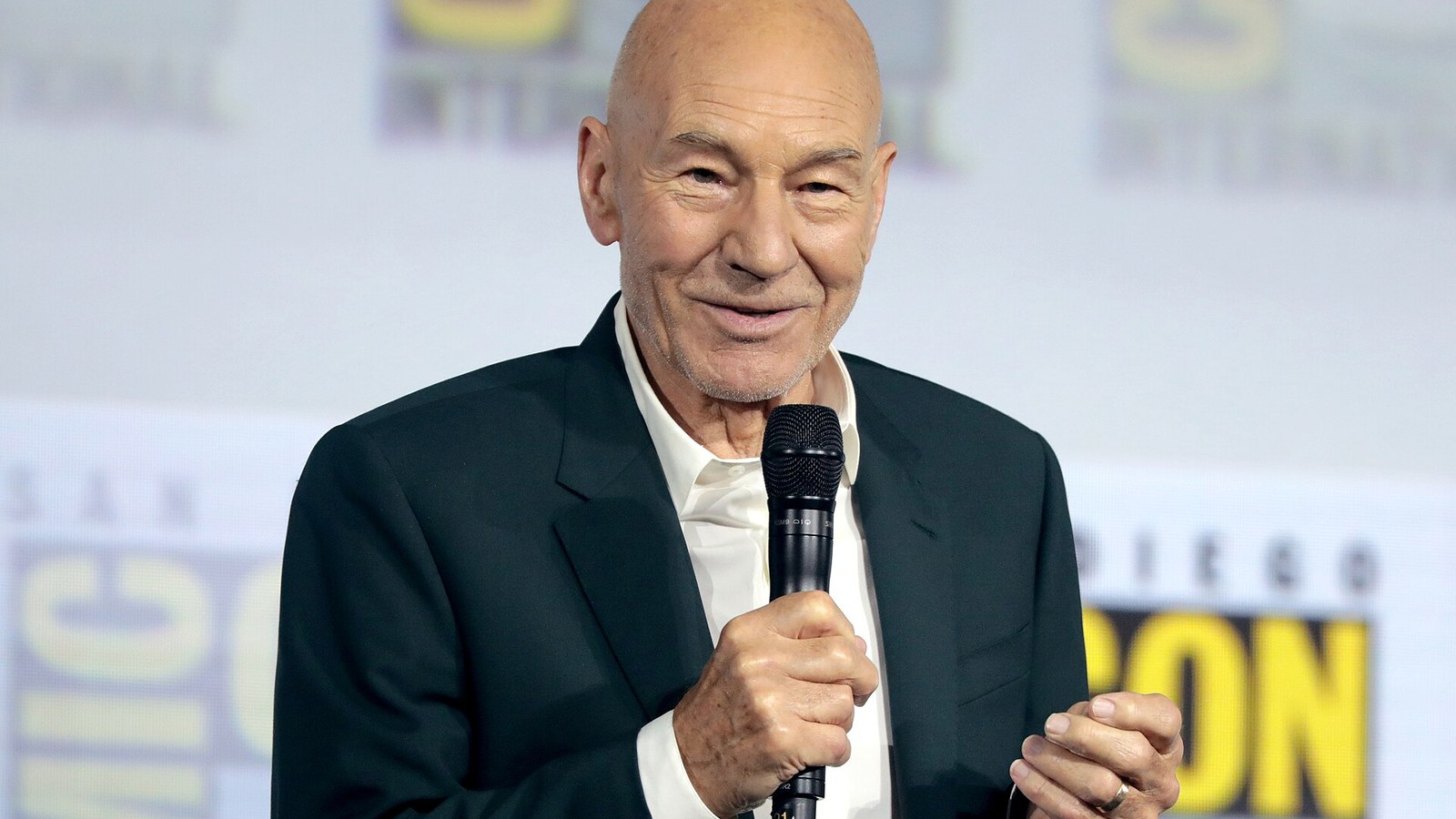 Patrick Stewart, en veste sombre et chemise blanche, parle dans un micro en souriant, devant un panneau jaune et blanc du Comic-Con.