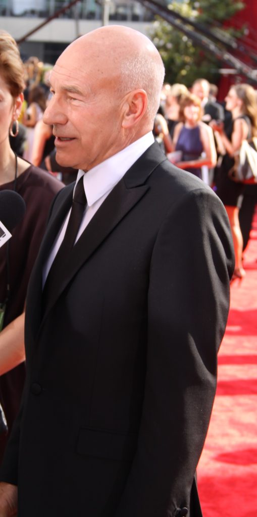 Patrick Stewart, en costume noir et cravate, sur le tapis rouge d'une cérémonie, de profil, devant une foule de photographes.