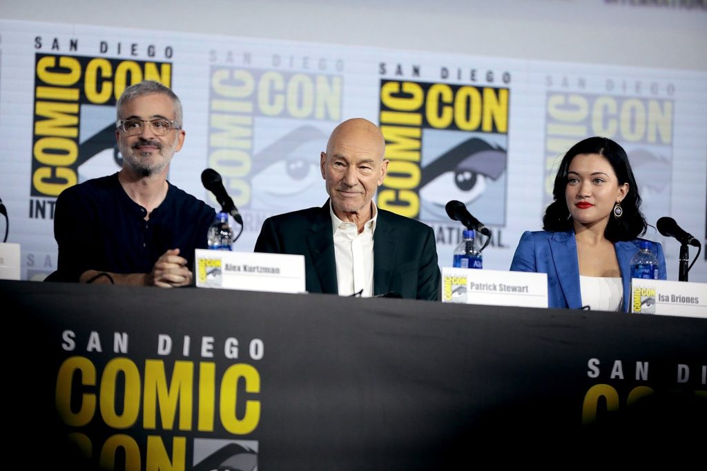 Trois personnes assises derrière une table, devant des micros et des affiches du Comic-Con de San Diego, dont Patrick Stewart au centre.