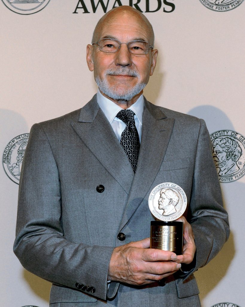 Patrick Stewart, barbe blanche taillée, en costume gris et cravate, tient une médaille dorée devant un mur portant le mot Awards.