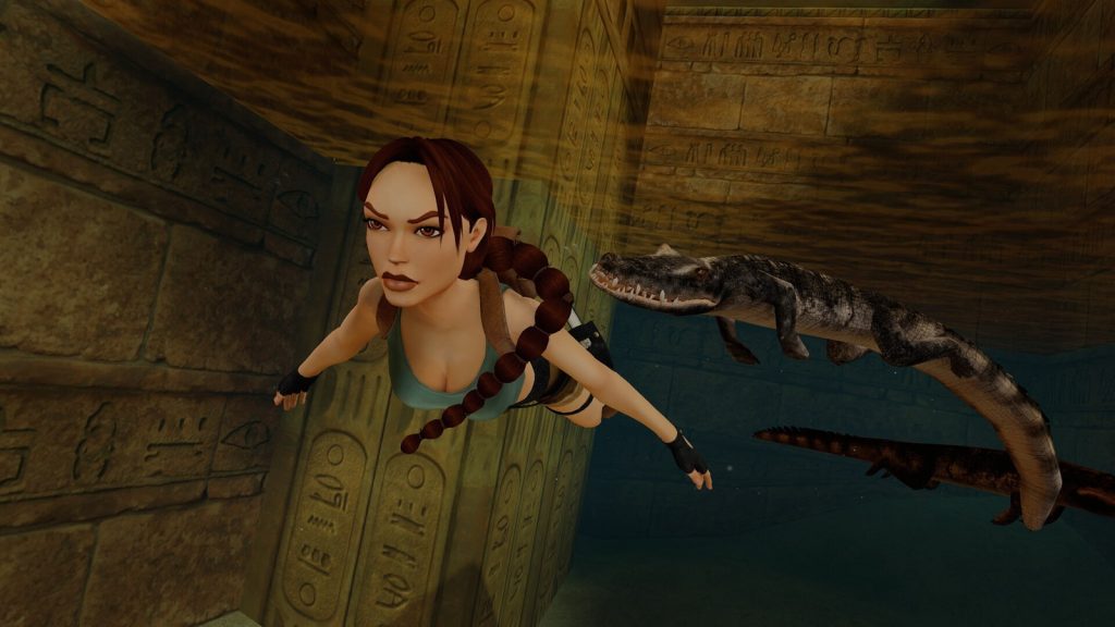 Lara Croft nage sous l'eau entre des murs gravés de hiéroglyphes, suivie par un crocodile.
