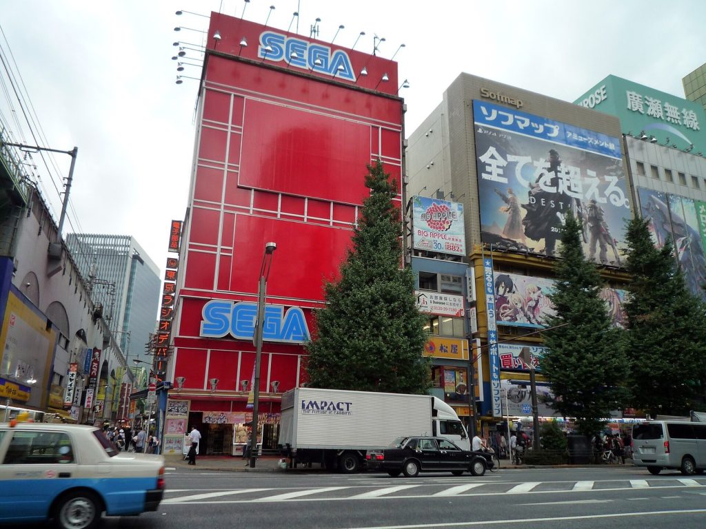 L'immeuble rouge de Sega à Akihabara, enseigne verticale au sommet, au milieu des panneaux publicitaires d'un carrefour de Tokyo.