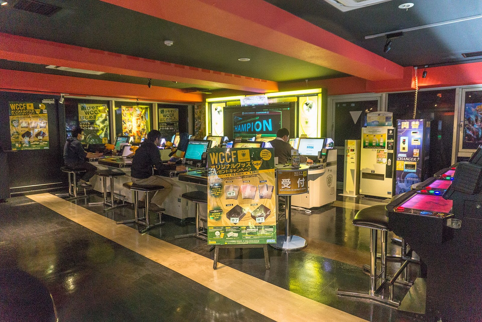 L'intérieur d'une salle d'arcade Sega, où des joueurs sont installés devant une rangée de bornes, sous un plafond orange.