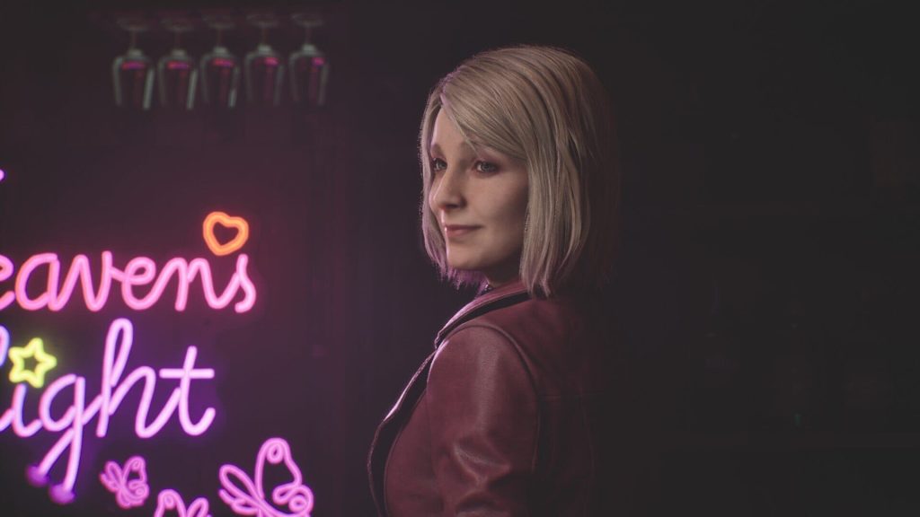 Une femme blonde en veste rouge sourit devant une enseigne au néon rose, dans une salle sombre.