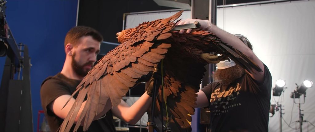 Deux artisans tiennent une grande marionnette d’aigle aux ailes de plumes brunes déployées, devant un fond bleu et des projecteurs.