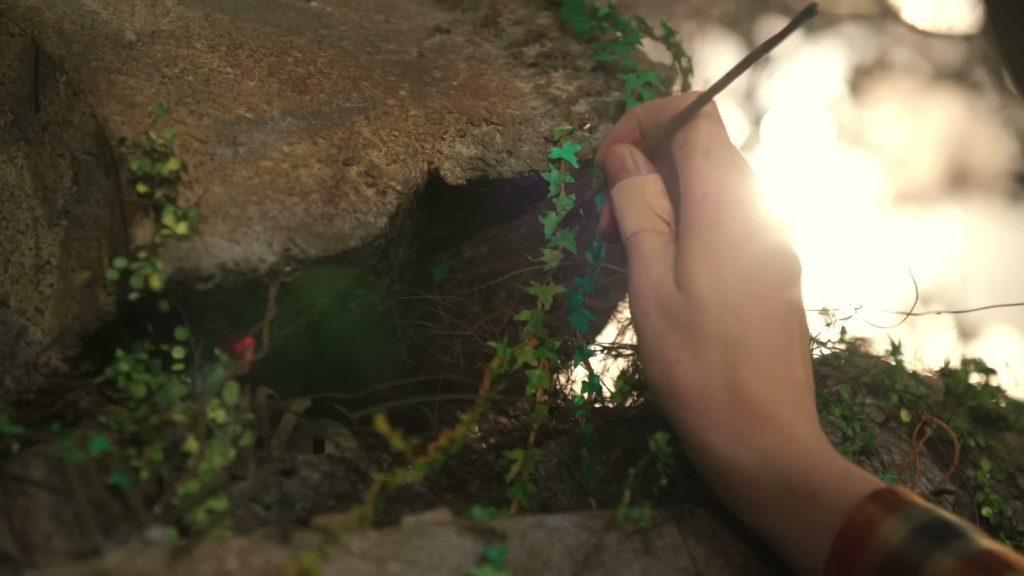 Une main tient un outil fin et dispose des brins de lierre miniature sur un rocher du décor, à contre-jour.