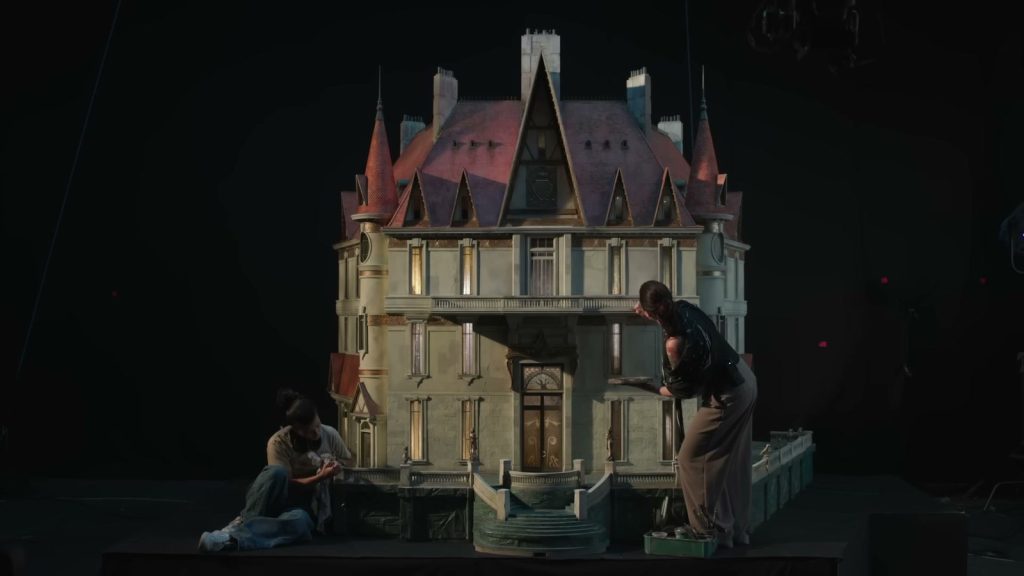 La maquette d’un manoir à tourelles, toit d’ardoise et fenêtres éclairées, dans un studio sombre, avec deux artisans au travail à sa base.