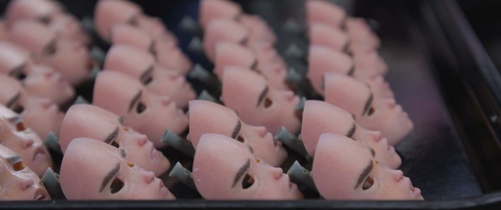 Des plateaux de petits visages de marionnette couleur chair, alignés en rangées et posés sur des tiges sombres.